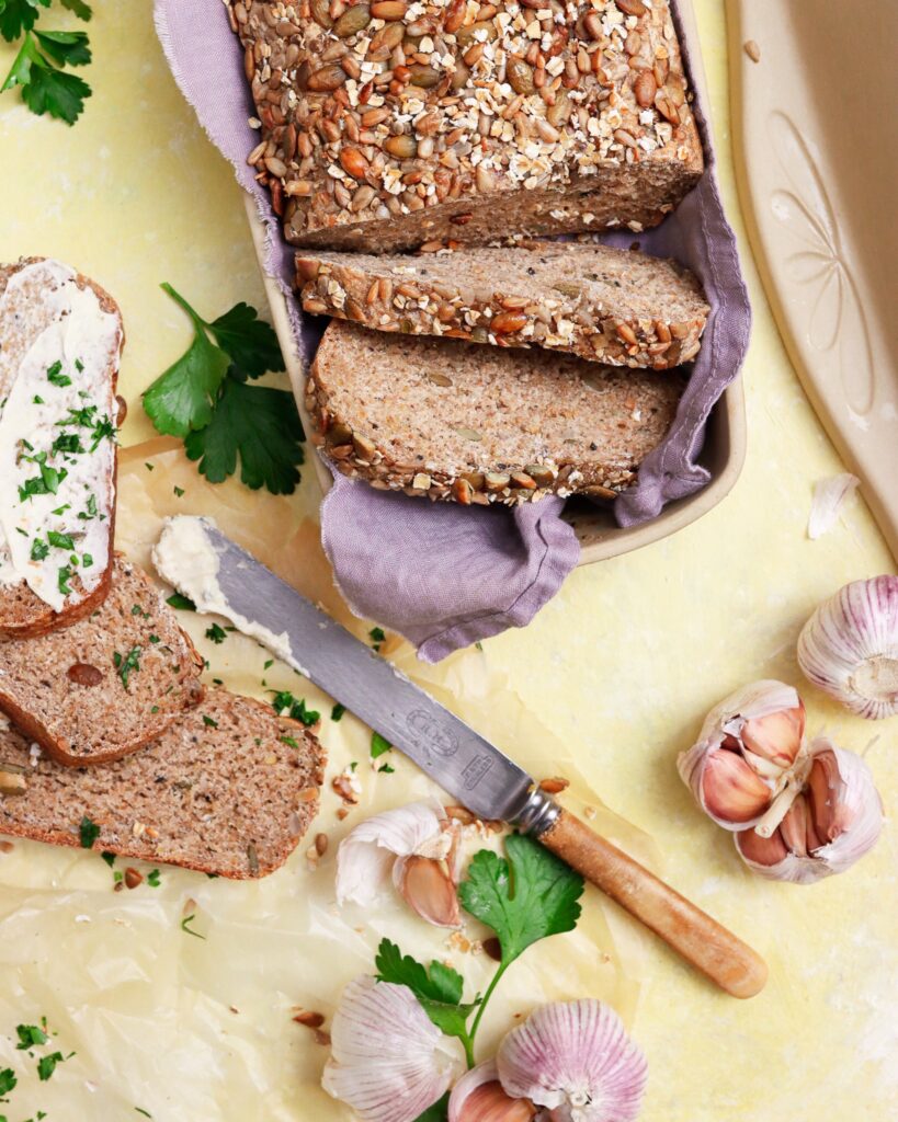 Fresh whole grain bread in artisan covered bread pan with garlic and spread on side.